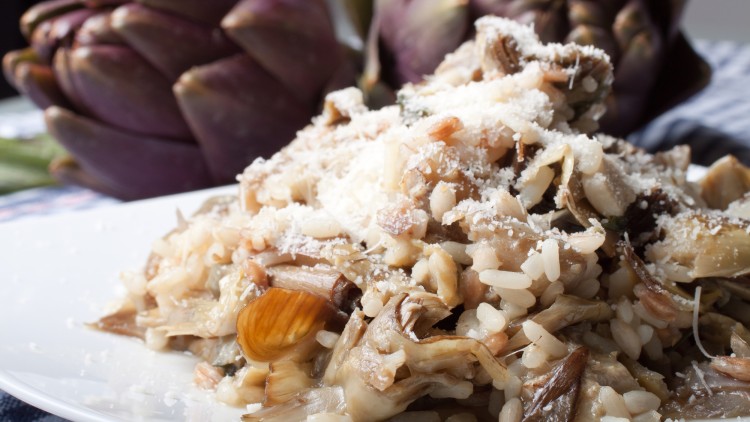 Ein Teller mit Risotto, Artischockenstücken und geriebenem Käse vor unscharfen lila Artischocken im Hintergrund.