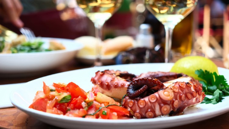 Ein Teller mit gebratenem Oktopus, Tomatensalat, Petersilie und einer Zitronenscheibe, im Hintergrund Weißwein und Salat.