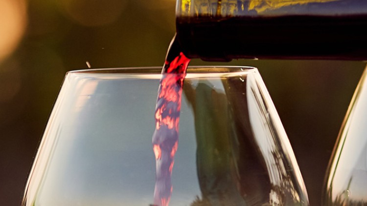Eine geöffnete Weinflasche gießt roten Wein in ein halbvolles Glas, daneben steht ein weiteres leeres Weinglas.