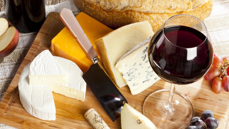 Ein Holzbrett mit verschiedenen Käsesorten, Brot, roten und blauen Weintrauben, Birne, Weinflasche, Weinkorken und Glas Rotwein.