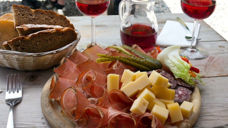 Holzbrett mit Käsewürfeln, Schinken, Salami, Gewürzgurken und Salat vor Brotkorb, Rotweingläsern und Karaffe.