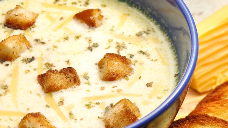 Eine cremige Suppe mit Croutons und Kräutern in einer blauen Schüssel, daneben gelber Käse und geröstete Brotscheiben.