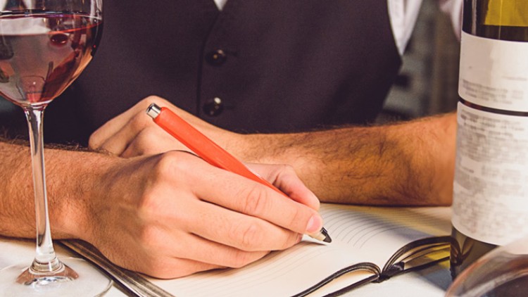 Person in weißem Hemd und Weste schreibt mit rotem Stift in ein Notizbuch, umgeben von zwei Weingläsern und Weinflaschen.