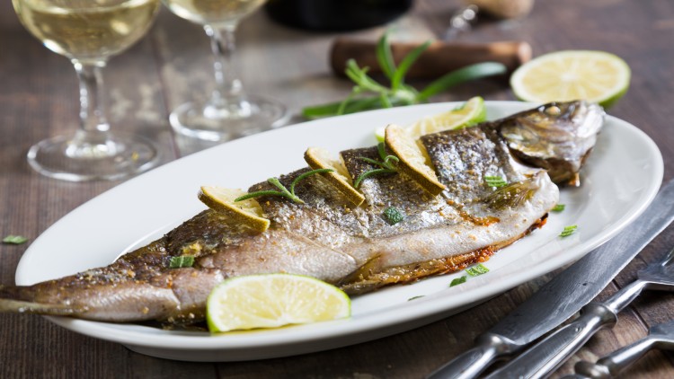 Gegrillter ganzer Fisch mit Zitronenscheiben und frischen Kräutern auf einem weißen Teller, daneben ein Besteck und Weingläser.