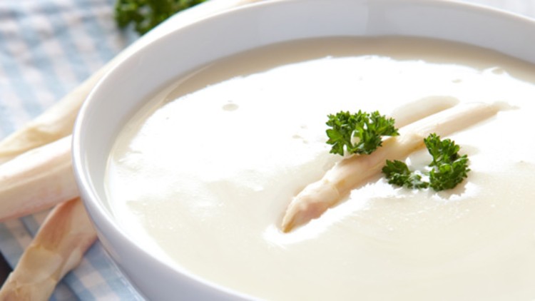 Eine hellweiße Spargelsuppe in einem weißen Teller, garniert mit Petersilie und zwei Spargelspitzen, auf kariertem Stoff.
