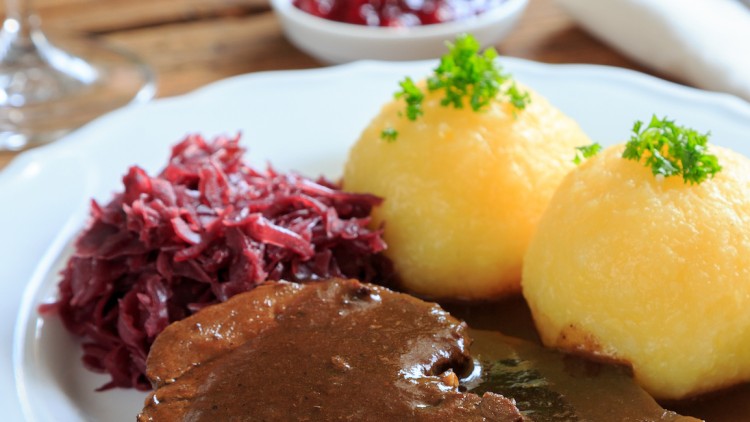 Auf einem weißen Teller befinden sich zwei Kartoffelklöße mit Petersilie, Rotkohl und ein Stück Braten mit dunkler Sauce.