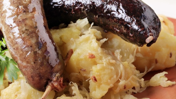 Zwei Bratwürste und eine Blutwurst auf Sauerkraut und Kartoffelstampf, garniert mit frischer Petersilie auf einem braunen Teller.