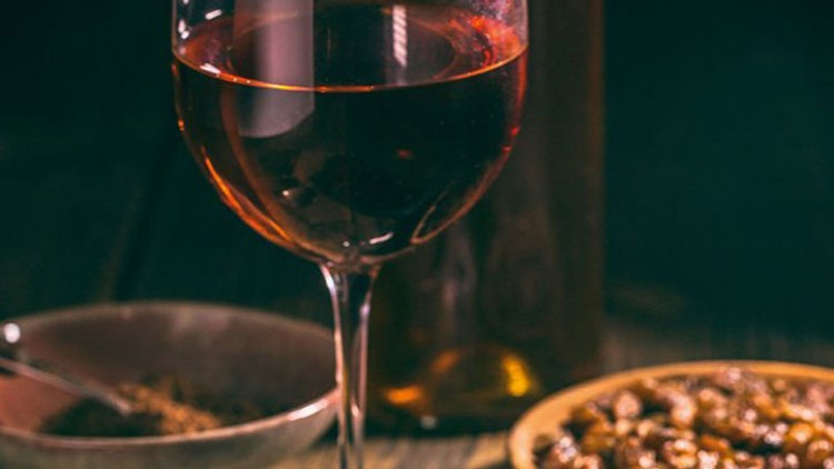 Ein Glas mit rotem Wein steht vor einer braunen Flasche, daneben Schalen mit Gewürzen und Nüssen auf einem Holztisch.