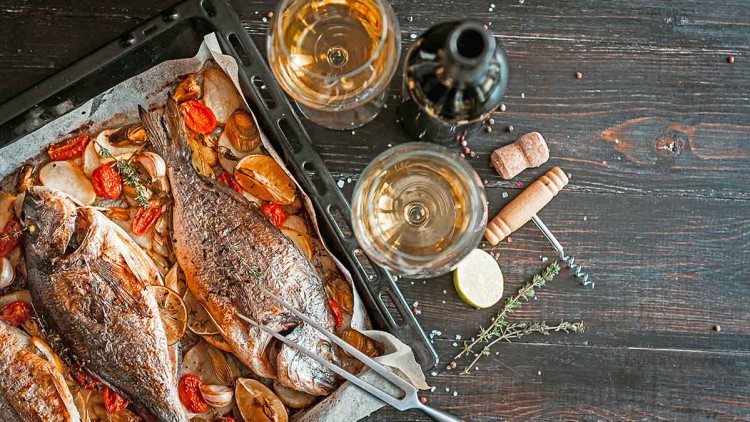 Zwei gegrillte Fische auf einem Backblech mit Tomaten, Zitronenscheiben und Kräutern, dazu zwei Weingläser und eine Weinflasche.