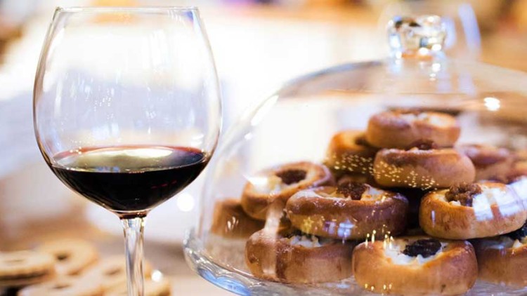 Ein Glas Rotwein und Zimtschnecken unter einer Glasglocke stehen auf einem hellen Tisch, daneben ein weiteres Weinglas.