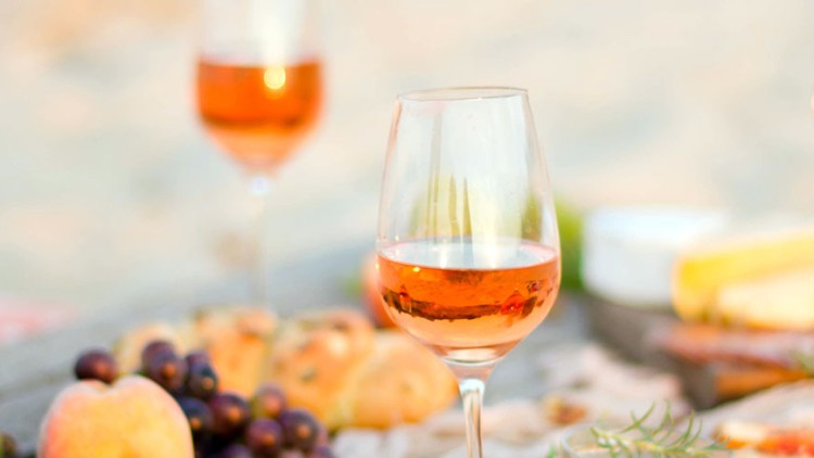 Drei Gläser Roséwein stehen auf einem Tisch mit Trauben, Brot, Pfirsichen und einer Käseplatte.