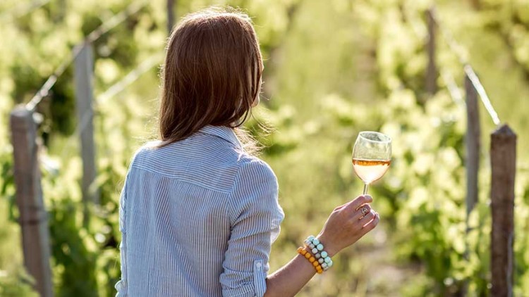 Eine Person von hinten in einem blau-weiß gestreiften Hemd hält ein Glas Weißwein und steht inmitten eines grünen Weinbergs.