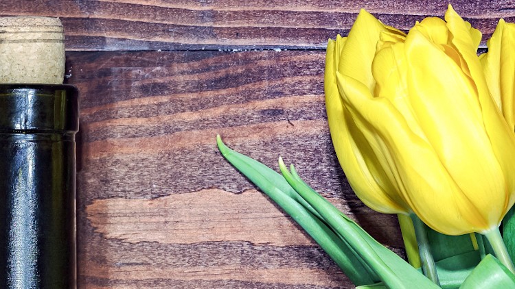 Gelbe Tulpen mit grünen Blättern liegen neben einer dunklen Weinflasche mit Korken auf einer Holzoberfläche.