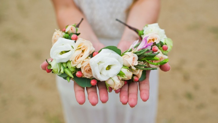 Zwei ausgestreckte Hände halten einen Blumenkranz aus weißen, beigefarbenen und rosafarbenen Blüten mit grünen Blättern.