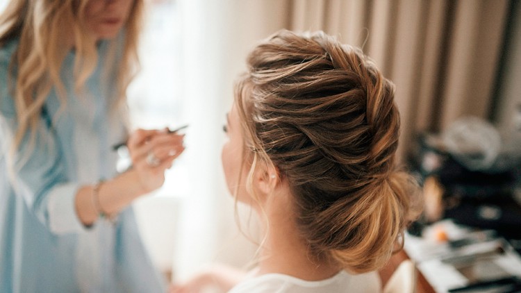 Person mit hochgestecktem Haar wird geschminkt, im Hintergrund unscharf Make-up-Utensilien auf einem Tisch erkennbar.