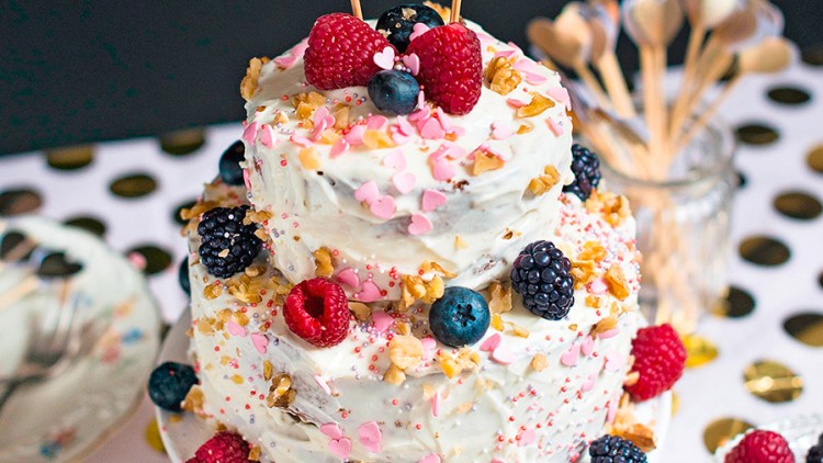 Zweistufige Torte mit weißem Überzug, Himbeeren, Blaubeeren, Brombeeren, Walnüssen und rosa Herzstreudekor, dekoriert mit Herzen.