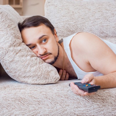 Eine Person liegt seitlich auf einem beigen Sofa, hält eine Fernbedienung in der Hand und lehnt an einem Kissen.