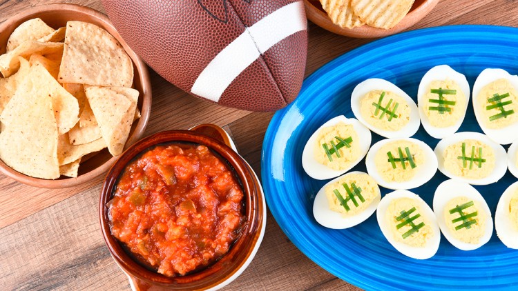 Ein brauner Football liegt neben Chips, Salsa-Dip, Kartoffelchips und gefüllten Eihälften mit grünem Garniturmuster auf Holztisch.