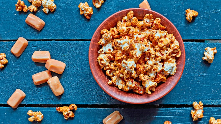 Eine braune Schale mit Karamell-Popcorn und lose verteilten Karamellbonbons auf einer blauen Holzoberfläche.