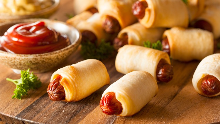 Blätterteig-Würstchen liegen auf einem Holztisch, daneben Petersilienzweige und Schalen mit Ketchup und Senf.