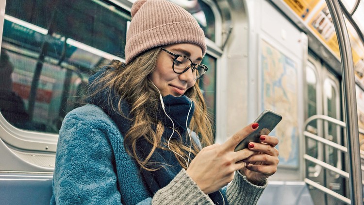 Person in blauer Jacke und rosa Mütze sitzt in der U-Bahn und hält ein Smartphone, weiße Kopfhörer sind angeschlossen.