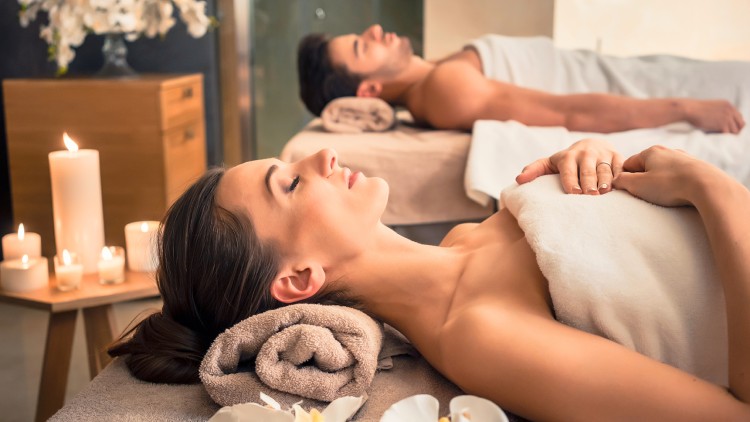 Zwei Personen liegen auf Massageliegen mit Handtüchern, Kerzen und weißen Blumen im Raum.
