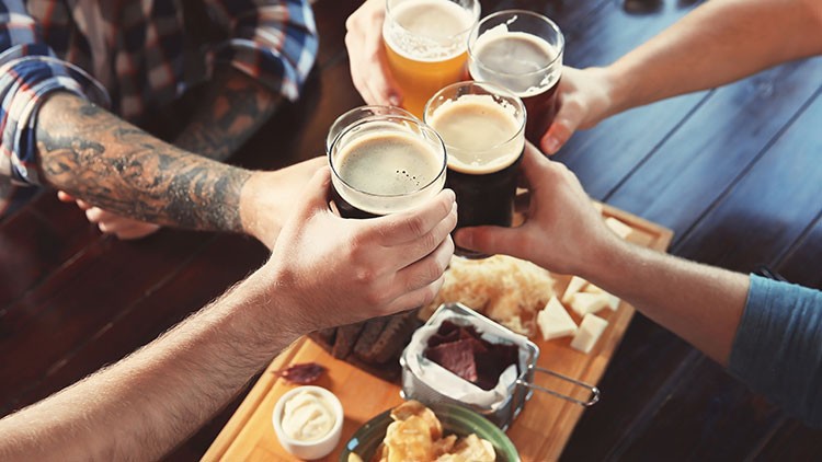 Vier Hände stoßen mit Gläsern Bier an, darunter ein Holztisch mit einer Platte aus Snacks wie Käse, Brot und Dips.