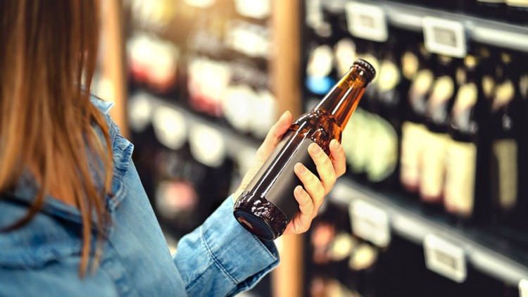 Person mit blauer Jeansjacke hält eine braune Glasflasche vor einem Regal mit weiteren Flaschen in einem Geschäft.