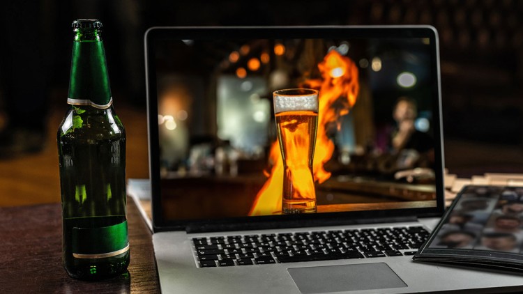 Ein aufgeklappter Laptop zeigt ein Glas Bier vor Flammen, daneben eine grüne Glasflasche und ein geschlossenes schwarzes Etui.