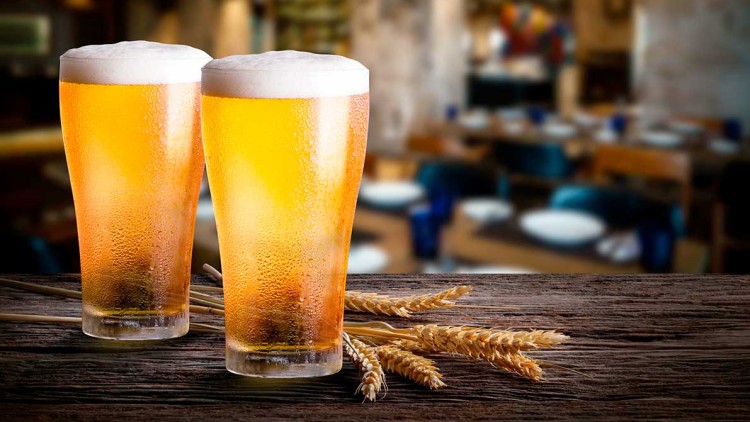 Zwei gefüllte Biergläser mit Schaum auf einem Holztisch, dekoriert mit Ähren, im Hintergrund unscharfe Restaurantumgebung.