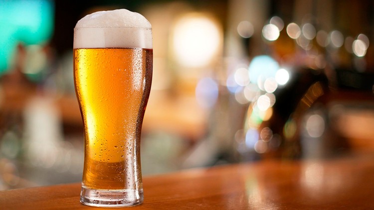 Ein hohes Glas mit goldenem Bier und Schaumkrone steht auf einer hölzernen Theke, im Hintergrund sind Lichter unscharf.