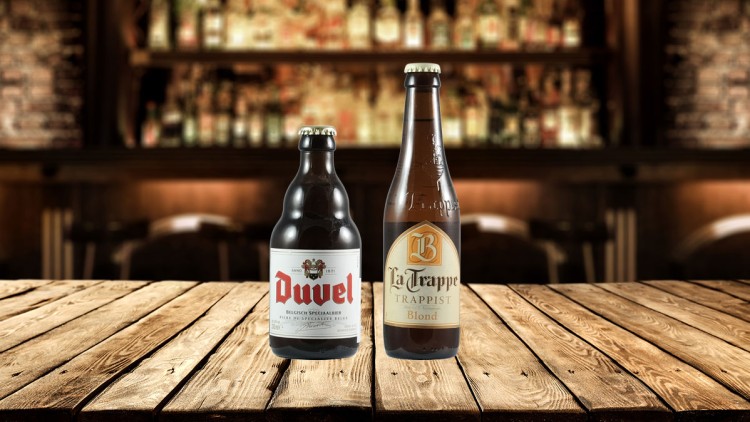 Zwei Bierflaschen, Duvel und La Trappe Blond, stehen auf einem Holztisch vor einer unscharfen Bar-Kulisse.
