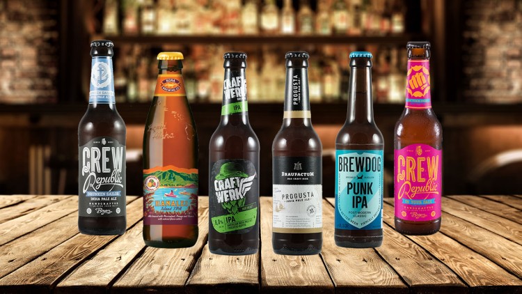 Sechs Flaschen Craft-Bier verschiedener Marken stehen auf einer Holzoberfläche vor einer unscharfen Bar-Umgebung.