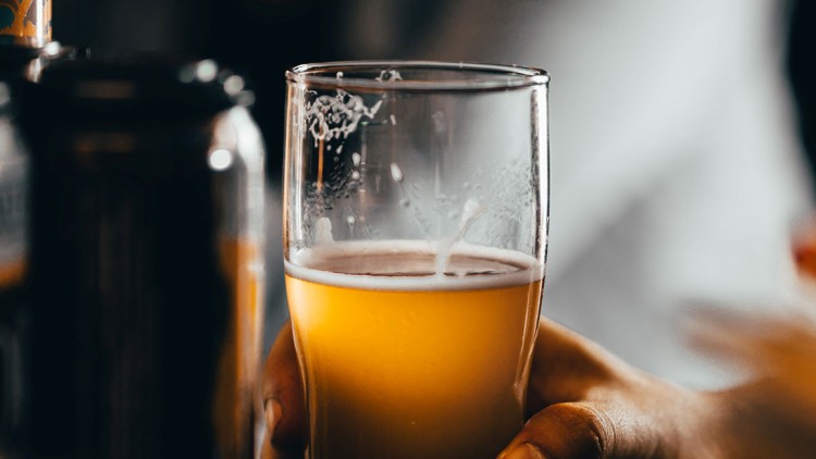 Ein halbvolles Glas mit goldgelbem Getränk wird von einer Hand gehalten, im Hintergrund sind unscharfe Dosen erkennbar.