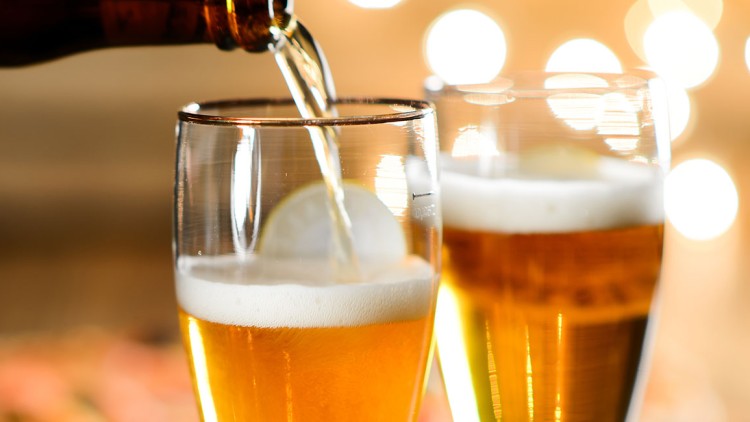 Eine Hand schenkt helles Bier aus einer Flasche in ein Glas mit Zitronenscheibe ein, weiteres gefülltes Glas im Hintergrund.