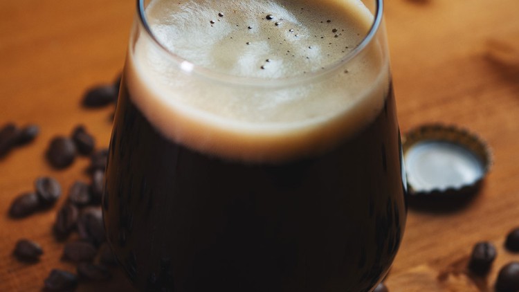 Ein gefülltes Glas mit dunklem Schaumgetränk steht auf einem Holztisch, umgeben von Kaffeebohnen und einem Kronkorken.