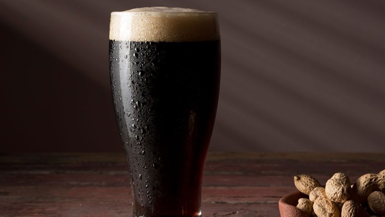 Ein Glas dunkles Bier mit Schaumkrone steht auf Holz, daneben liegt eine Schale mit Erdnüssen in Schalen.