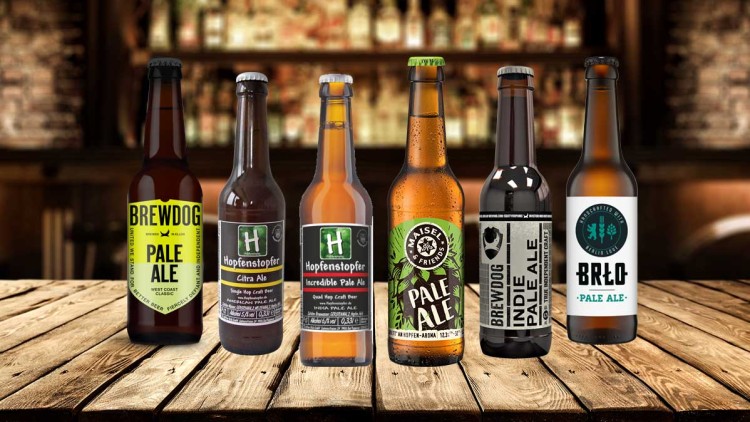 Sechs verschiedene Pale Ale Bierflaschen stehen nebeneinander auf einem Holztisch, unscharfer Hintergrund mit Barregalen.