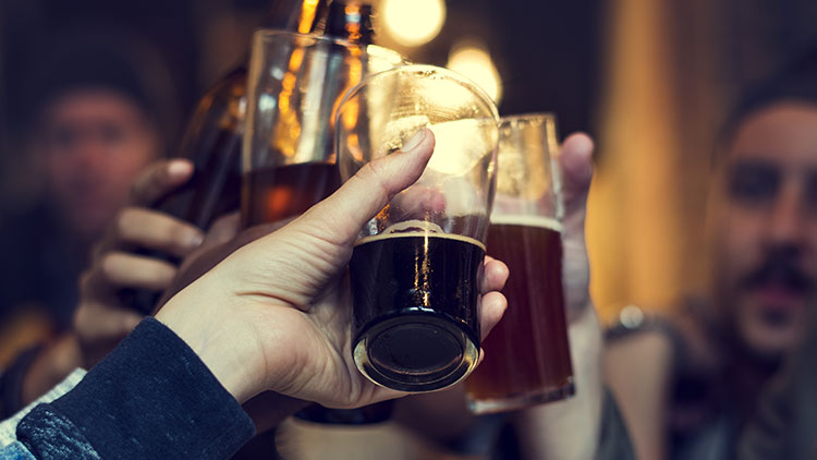 Mehrere Hände halten gefüllte Biergläser in die Höhe, Hintergrund unscharf und warm beleuchtet.
