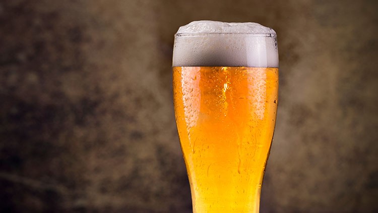 Ein gefülltes Bierglas mit goldenem Getränk und weißer Schaumkrone vor unscharfem braunem Hintergrund.