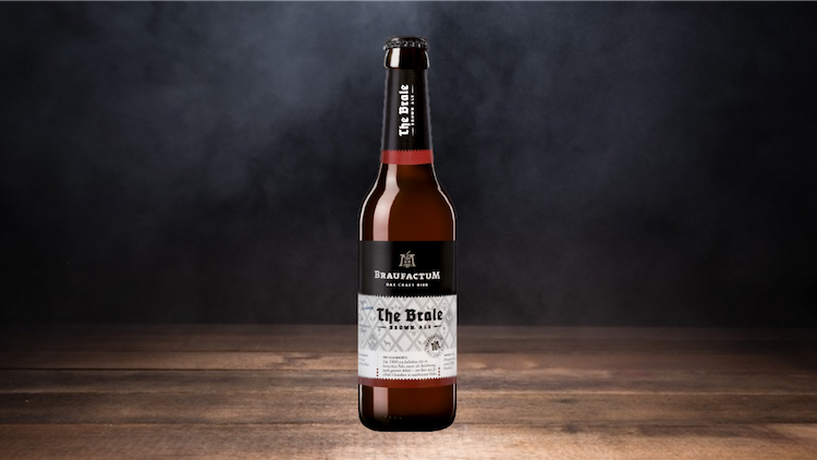 Braune Bierflasche mit der Aufschrift „BraufactuM, The Brale, Brown Ale“ auf Holzuntergrund und dunklem, nebligem Hintergrund.