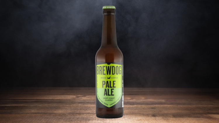 Eine braune Glasflasche mit grünem Etikett und der Aufschrift „BREWDOG PALE ALE“ steht auf einem Holztisch vor dunklem Hintergrund.
