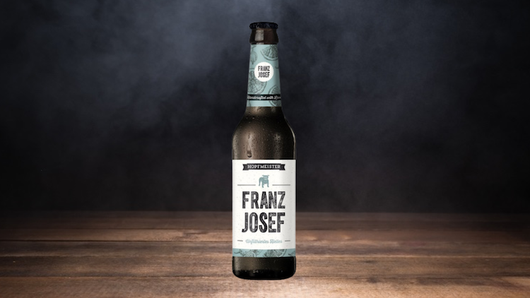 Braune Bierflasche mit Etikett „Hopfmeister Franz Josef“ steht vor dunklem Hintergrund auf einer Holzoberfläche.