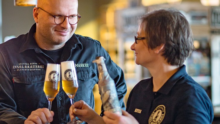 Zwei Personen stoßen mit Biergläsern an, eine Person hält zusätzlich eine Flasche, im Hintergrund warme Beleuchtung.