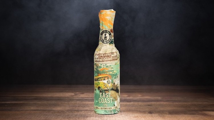 Eine in bunt bedrucktes Papier gewickelte Bierflasche auf einem dunklen Holzuntergrund mit schwarzem Rauch im Hintergrund.