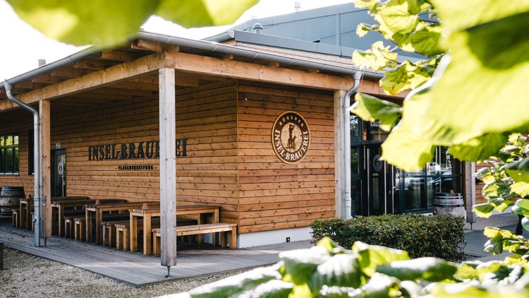 Holzgebäude mit der Aufschrift „Rügener Insel-Brauerei Flaschenreifung“ und Tischen, umgeben von Pflanzen und einem Kiesweg.