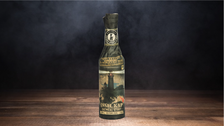 Eine dunkle Glasflasche mit Papierhülle, darauf ein Leuchtturmmotiv und der Text „INSEL KAP OATMEAL STOUT“.