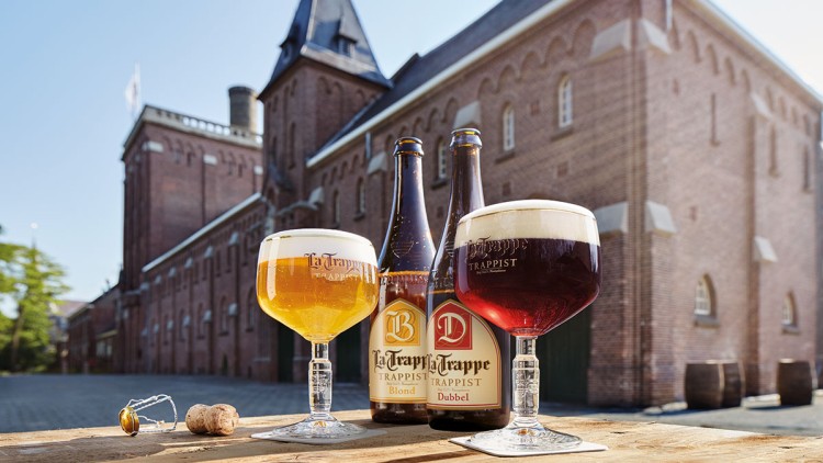 Zwei Biergläser mit hellem und dunklem Bier stehen vor zwei passenden Flaschen vor einem roten Backsteingebäude.