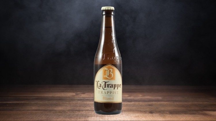 Braune Bierflasche mit Etikett „La Trappe Trappist Blond“ auf hölzerner Oberfläche, dunkler Hintergrund mit leichtem Nebel.