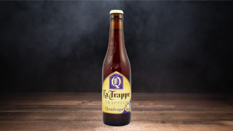 Braune Bierflasche mit gelbem Etikett, Aufschrift „La Trappe Trappist Quadrupel“ auf Holzuntergrund vor dunklem Hintergrund.